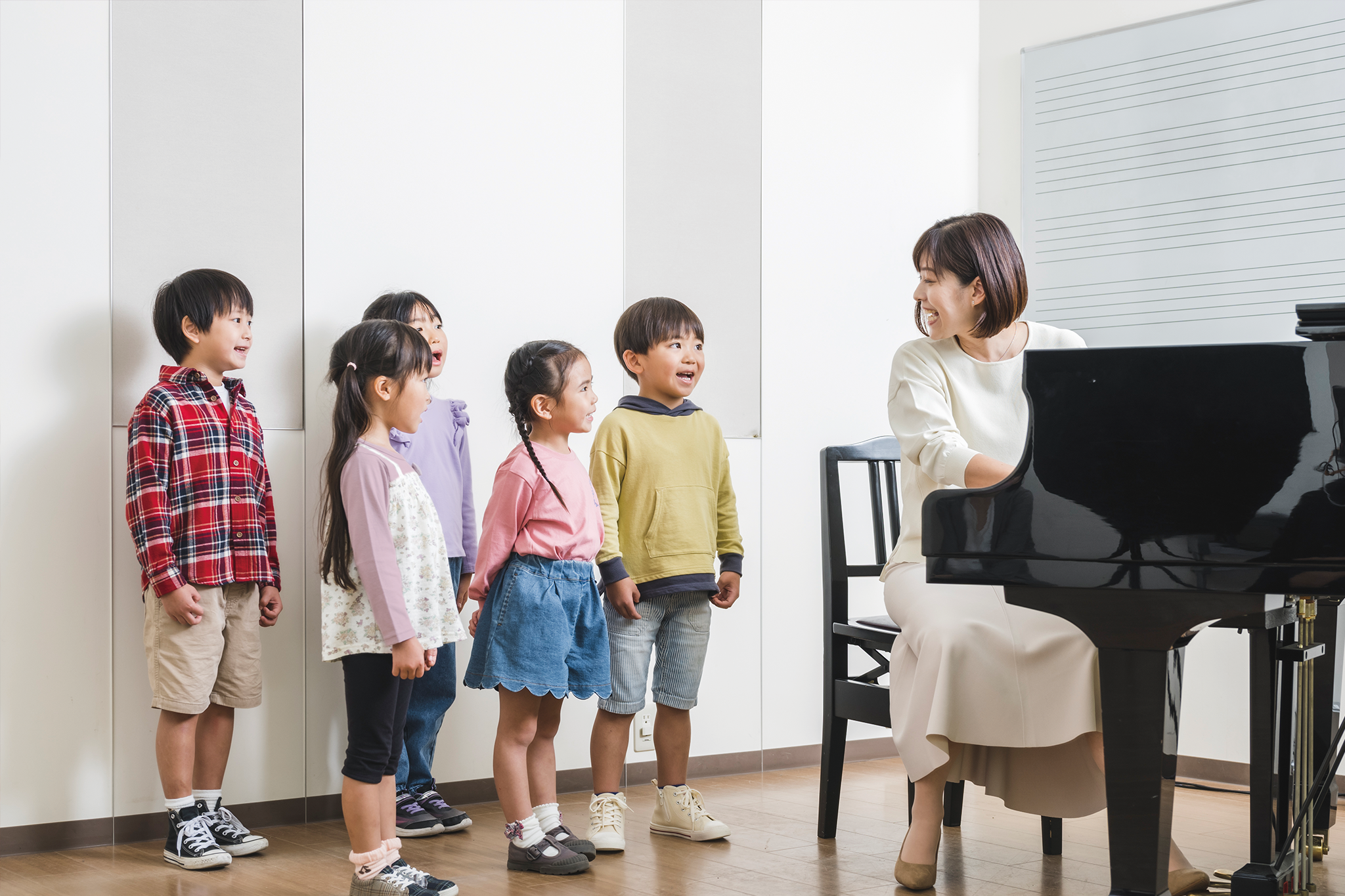 A cheerful music teacher engages a group of children aged 4 to 5.5 as they sing together beside a grand piano in a lively classroom setting with a whiteboard behind them